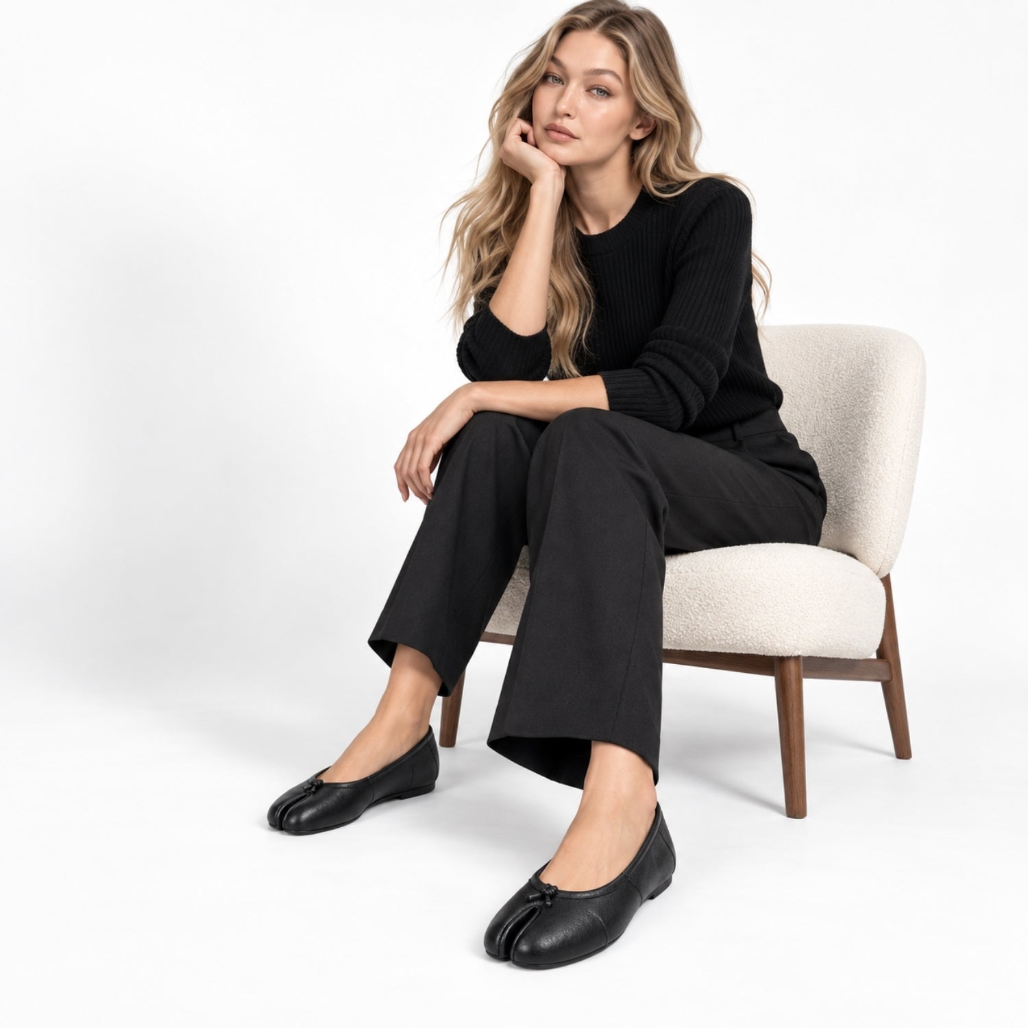 A woman sits on a cream upholstered chair wearing a black ribbed long-sleeve top, black wide-leg cropped pants, and black tab-flat shoes against a clean white studio background.