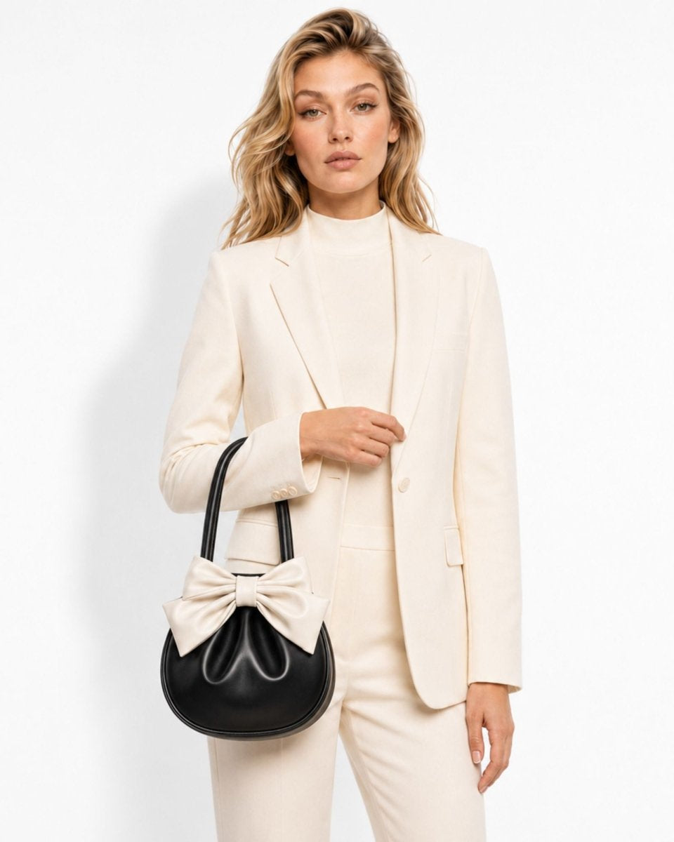 A woman wearing a cream-colored blazer and matching pants holds a black crescent-shaped handbag with a large cream bow detail, set against a plain white studio background.