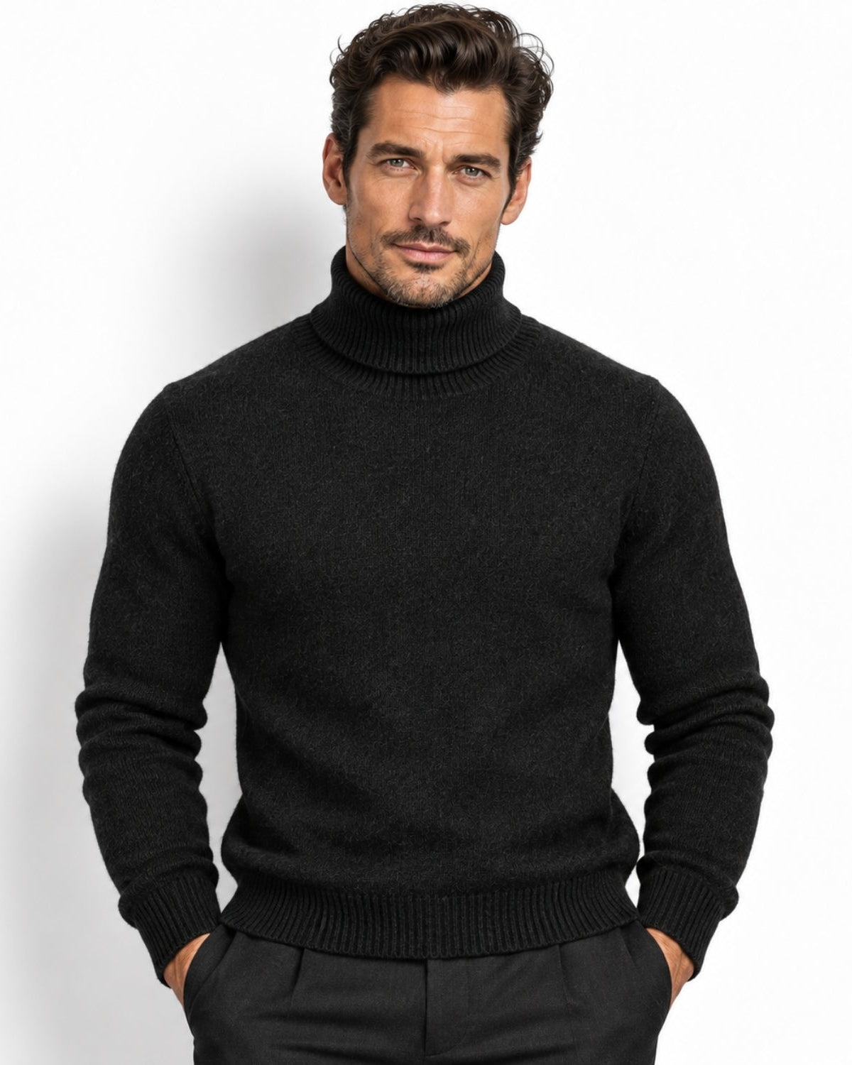 A man wearing a black knit turtleneck sweater and dark trousers, standing against a plain white studio background.