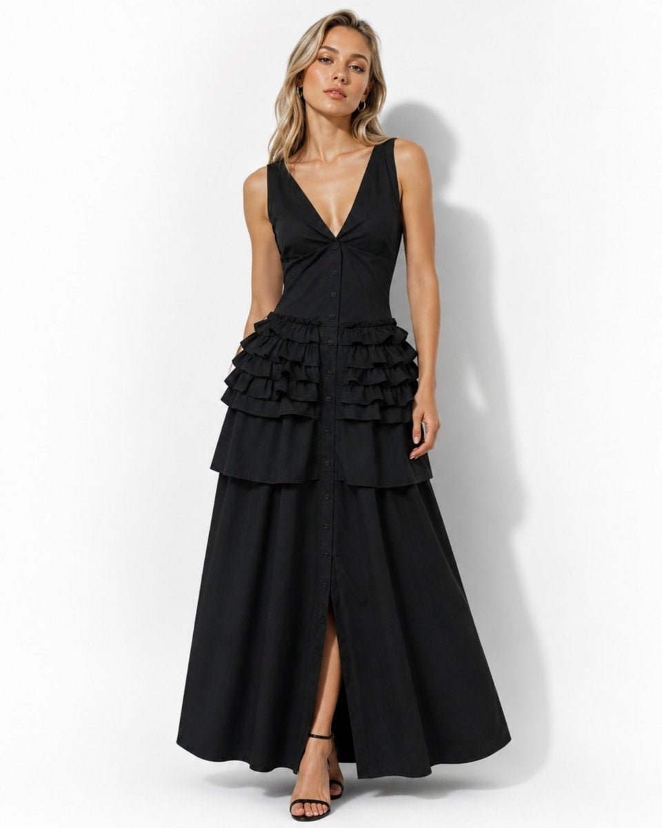 A woman modeling a sleeveless black maxi dress with a deep V-neckline, structured corset-style bodice, tiered ruffle waist, and a front slit, photographed against a plain light gray studio background.