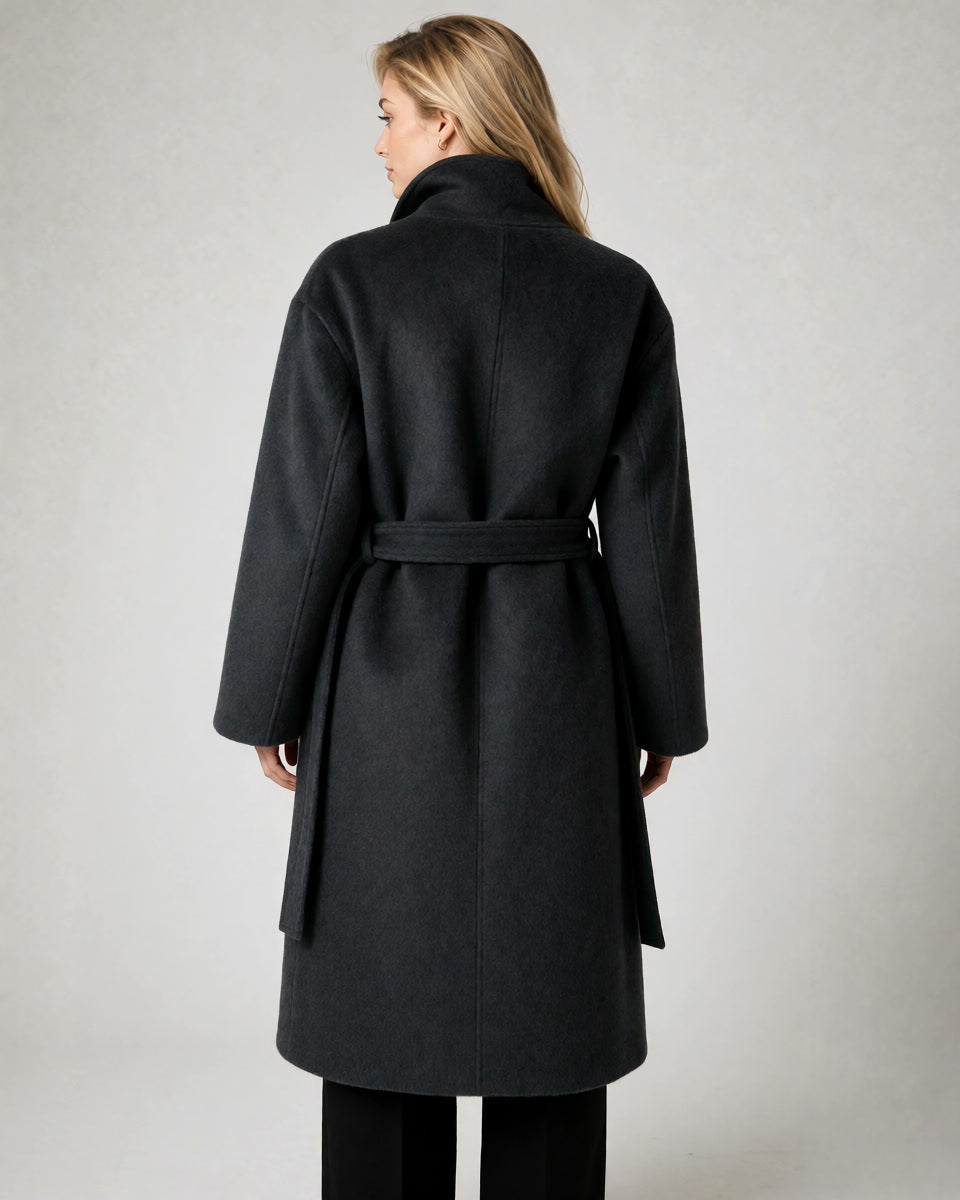 A woman viewed from the back wearing a dark charcoal gray belted coat with a high collar, paired with black pants, against a neutral light gray background.