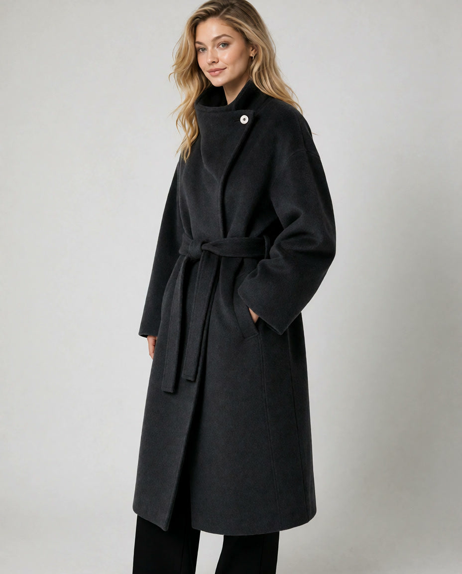A woman modeling a long black wrap coat with a self-tie belt and a single decorative button at the collar, photographed against a plain light gray studio background.