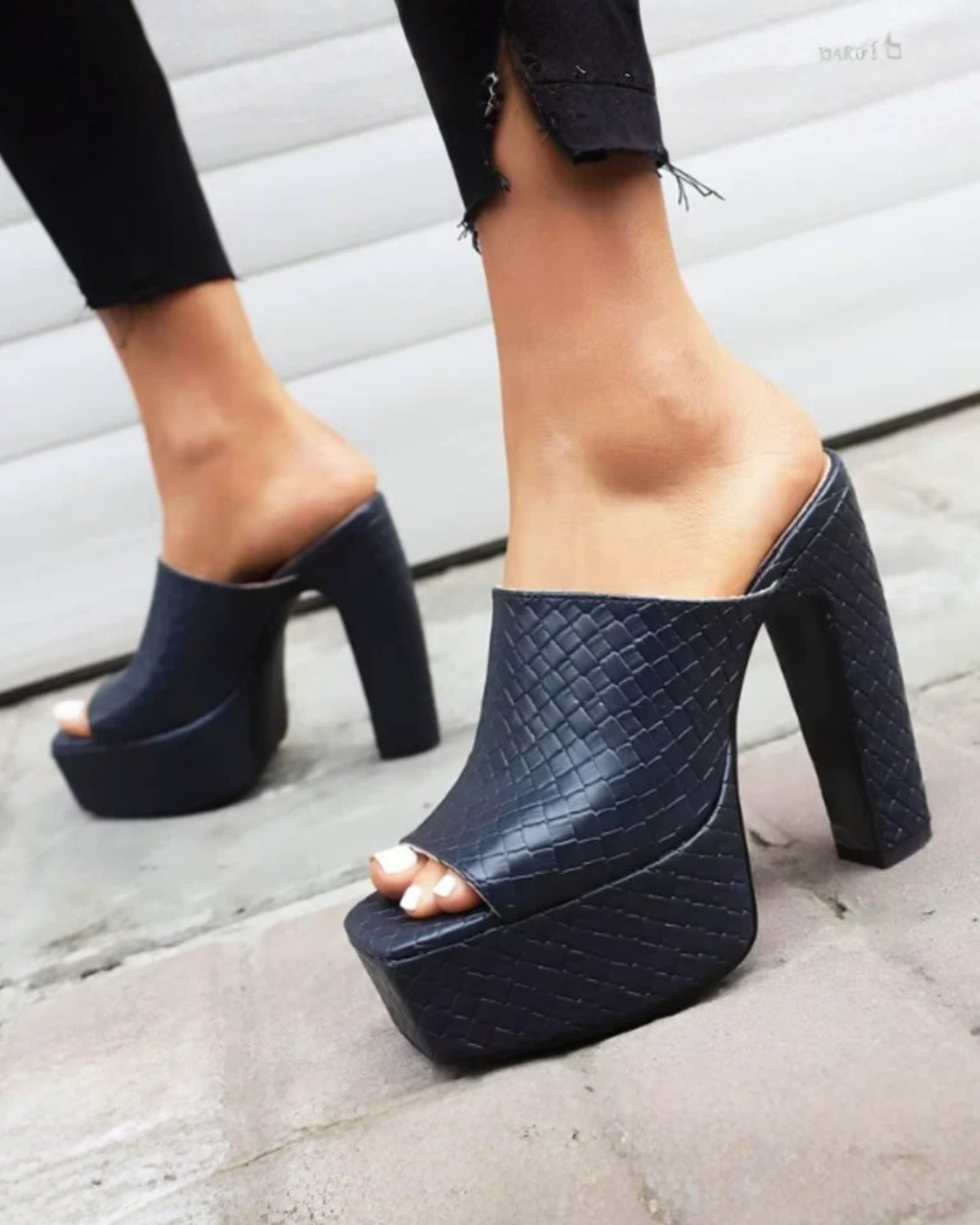 A pair of women's black platform mule heels with a textured crocodile-embossed pattern, shown on a model's feet against a gray concrete and white paneled wall background.