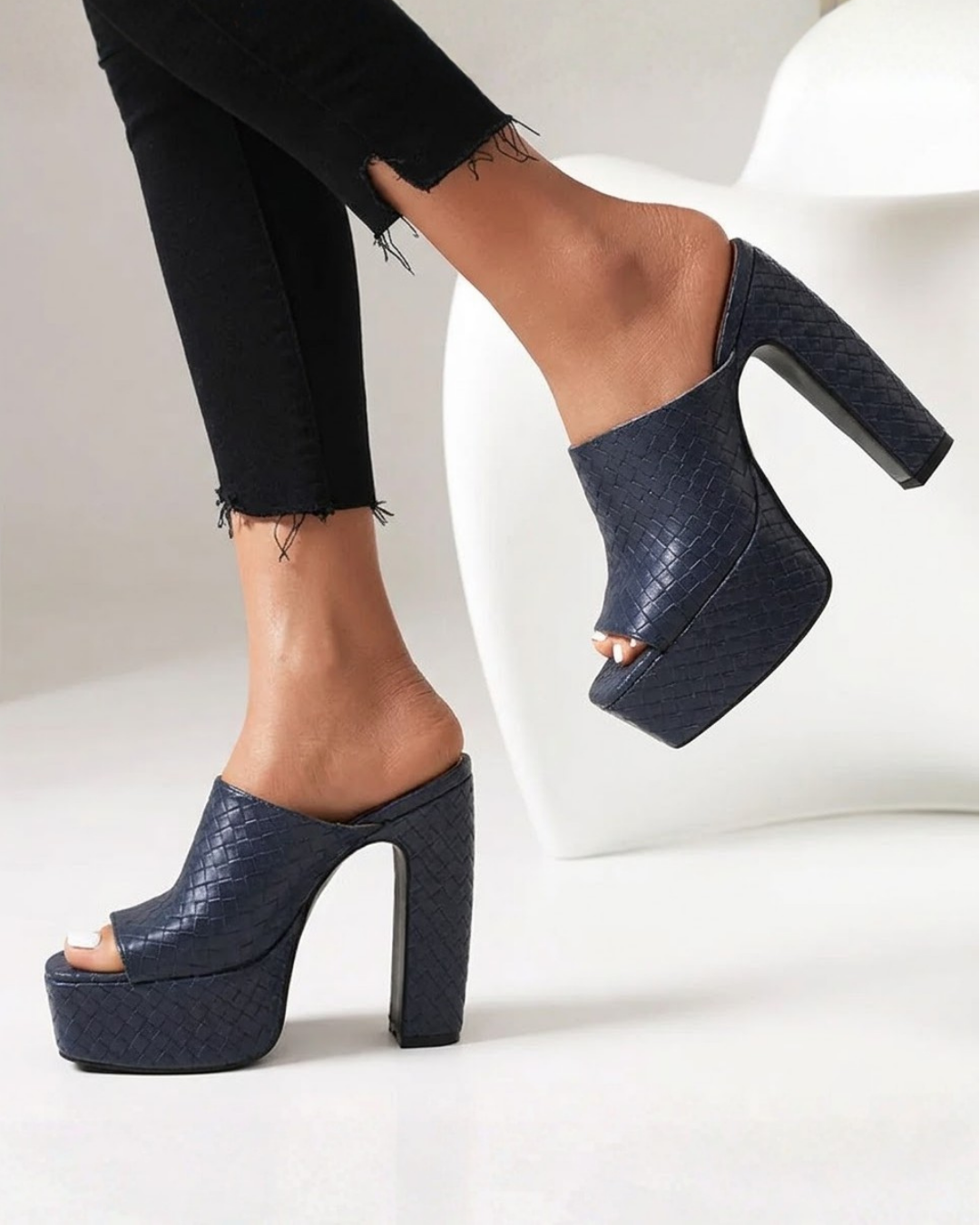 A pair of dark navy blue platform mule heels with a textured python-like pattern, shown on a model wearing black frayed-hem jeans against a clean white studio background.
