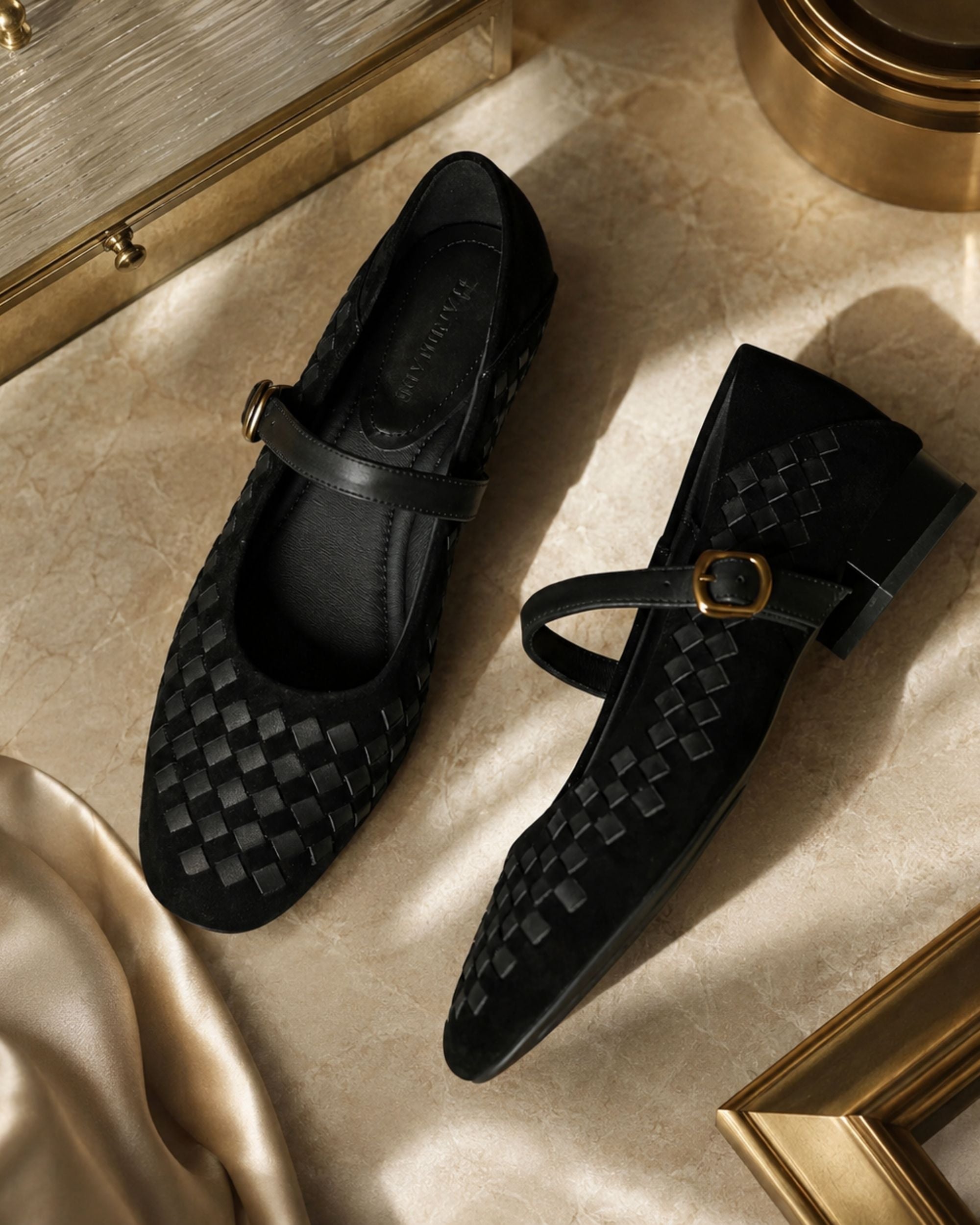 A pair of black woven Mary Jane flats with a single strap and gold-tone buckle, styled on a beige marble surface with gold-toned decorative props.