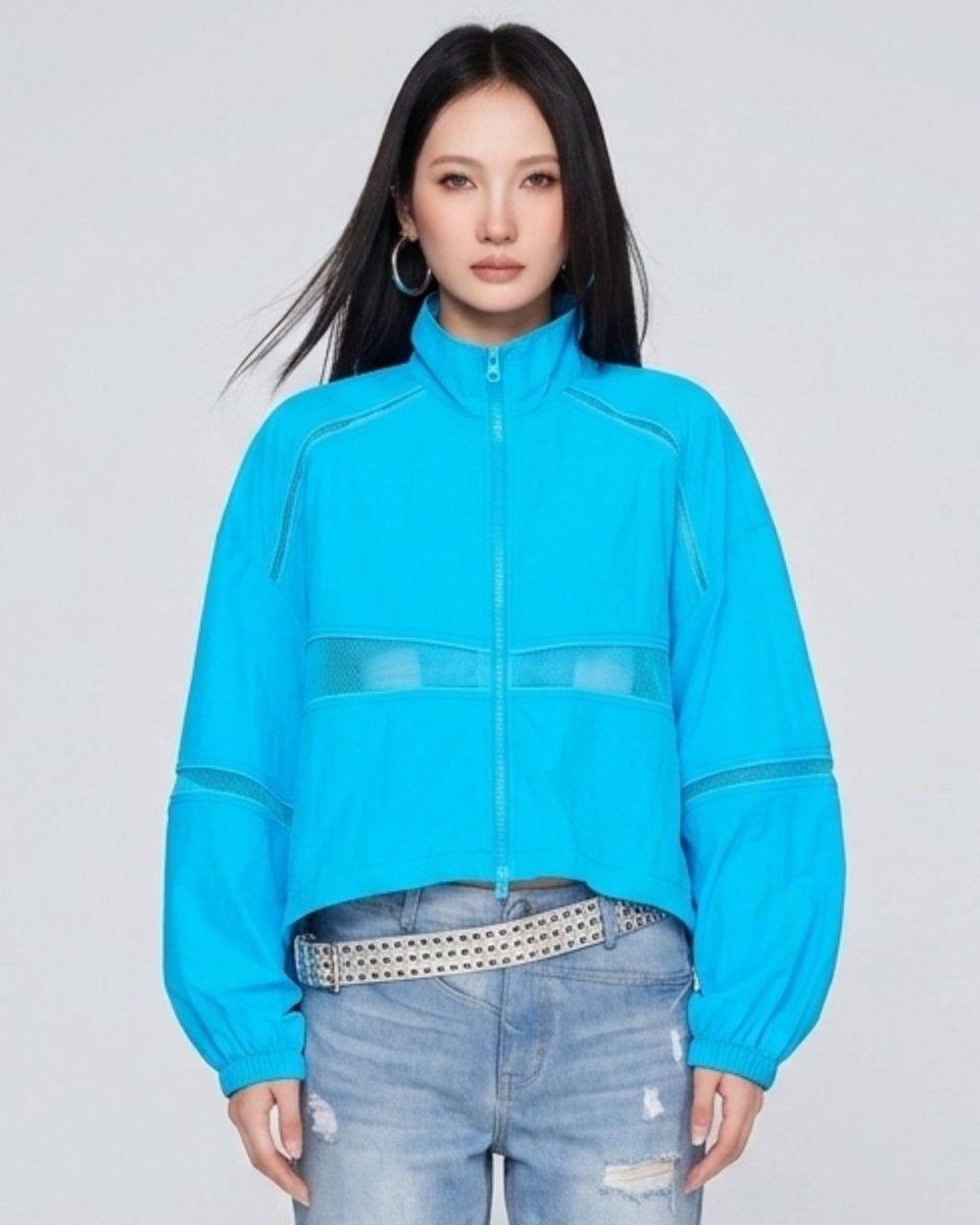Woman wearing Kouvr bright blue cropped windbreaker track jacket with sheer mesh panels, front studio view.