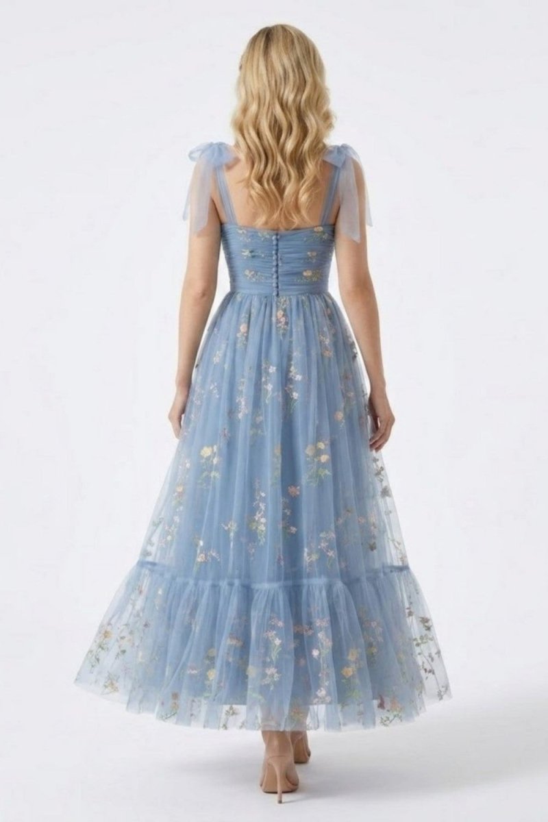 A woman viewed from behind wearing a light blue tulle maxi dress with floral embroidery and sheer flutter sleeves, standing against a plain light gray studio background.