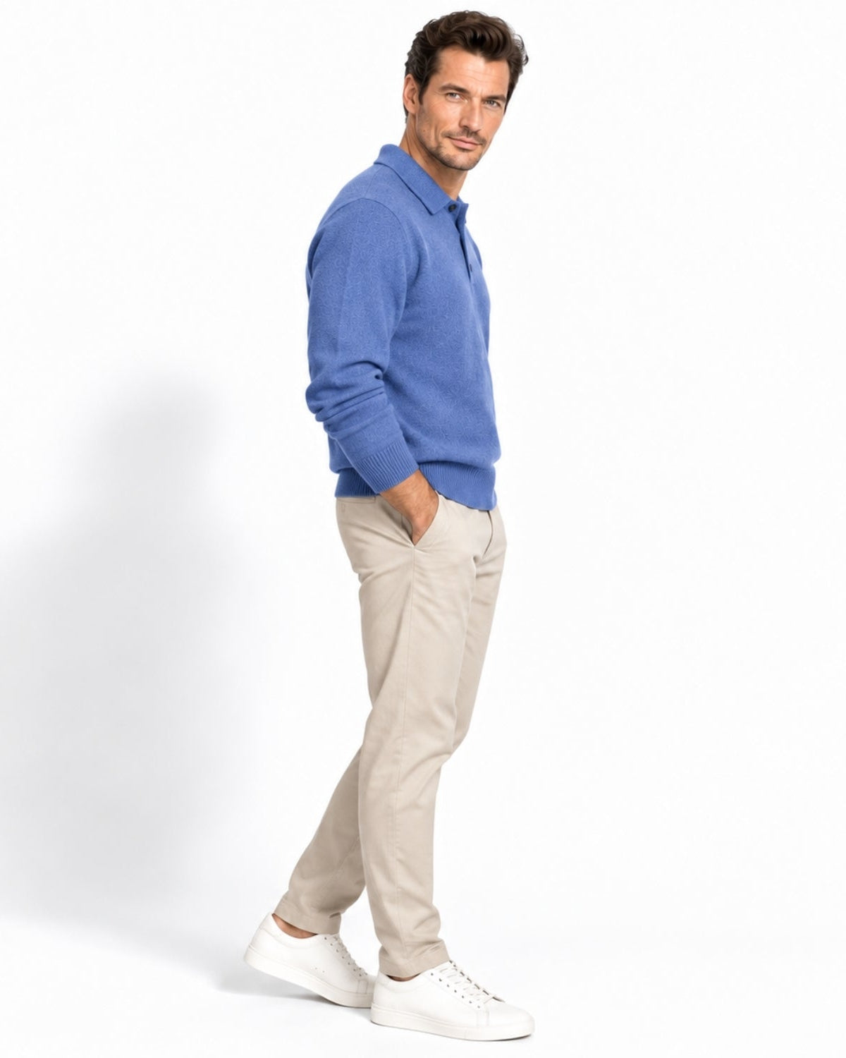 A man wearing a blue long-sleeve polo shirt, beige trousers, and white sneakers, standing in profile against a clean white studio background.