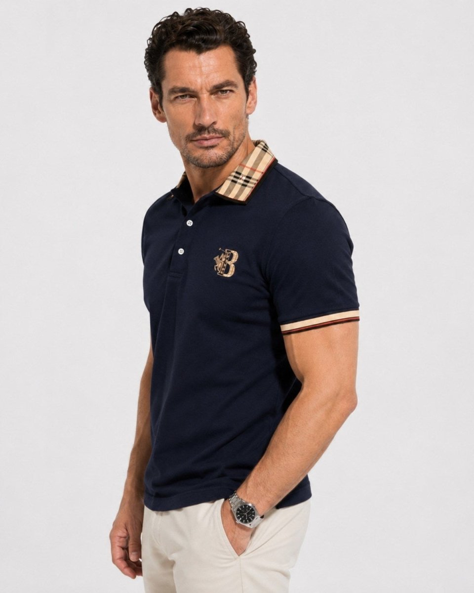 A man wearing a navy blue polo shirt with a beige and brown checkered collar, a gold embroidered crest on the chest, and striped trim on the sleeve cuffs, paired with off-white trousers, against a light gray studio background.