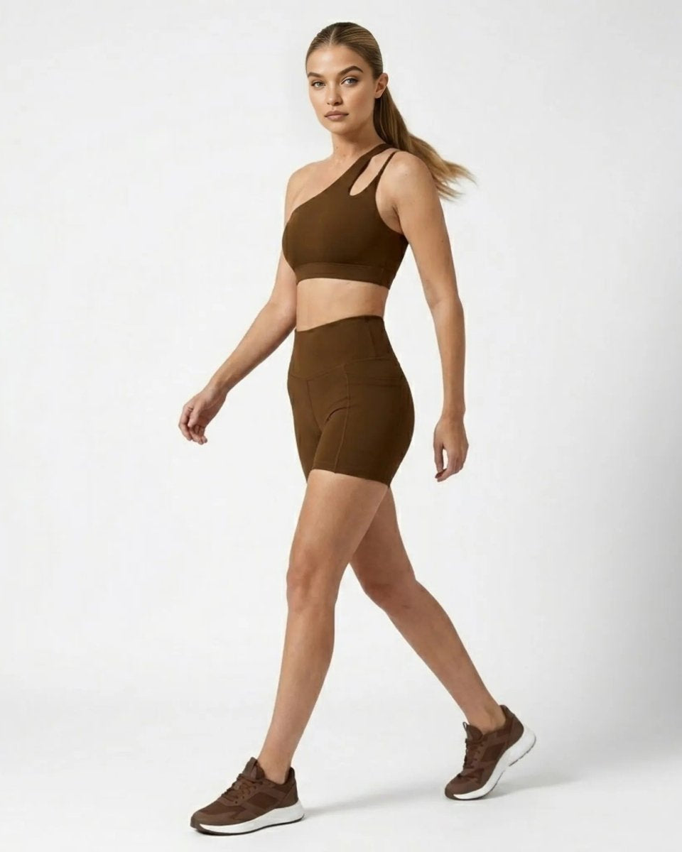 Profile view of woman walking in brown asymmetrical matching gym outfit with side pockets