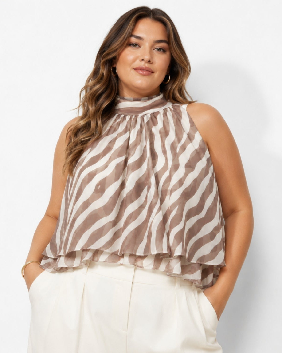 A woman wearing a sleeveless high-neck blouse with a brown and cream wavy stripe pattern, paired with white trousers, against a plain white studio background.