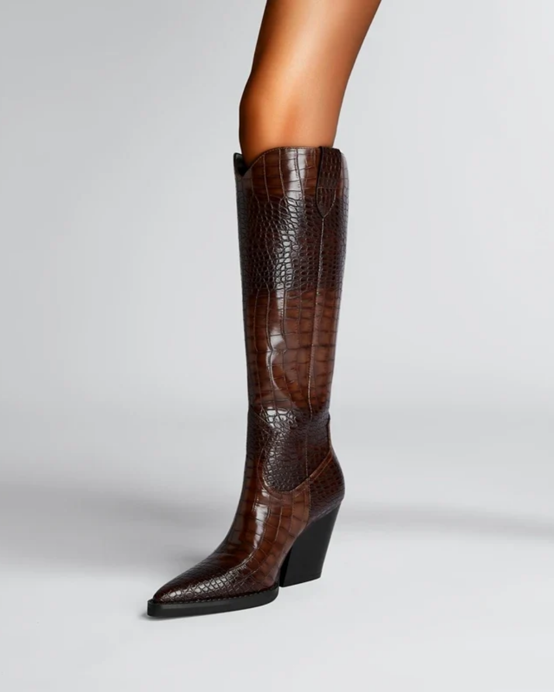 A knee-high brown boot with a crocodile-embossed texture, featuring a pointed toe and a black wedge heel, shown on a model's leg against a light gray background.