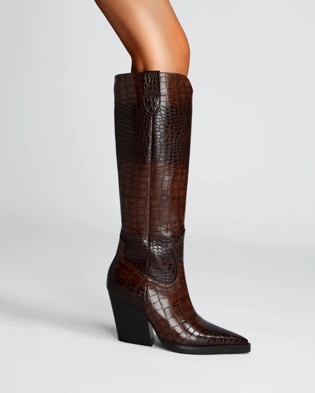 A dark brown knee-high boot with an embossed crocodile pattern, featuring a pointed toe and a chunky black block heel, shown on a model's leg against a light gray background.