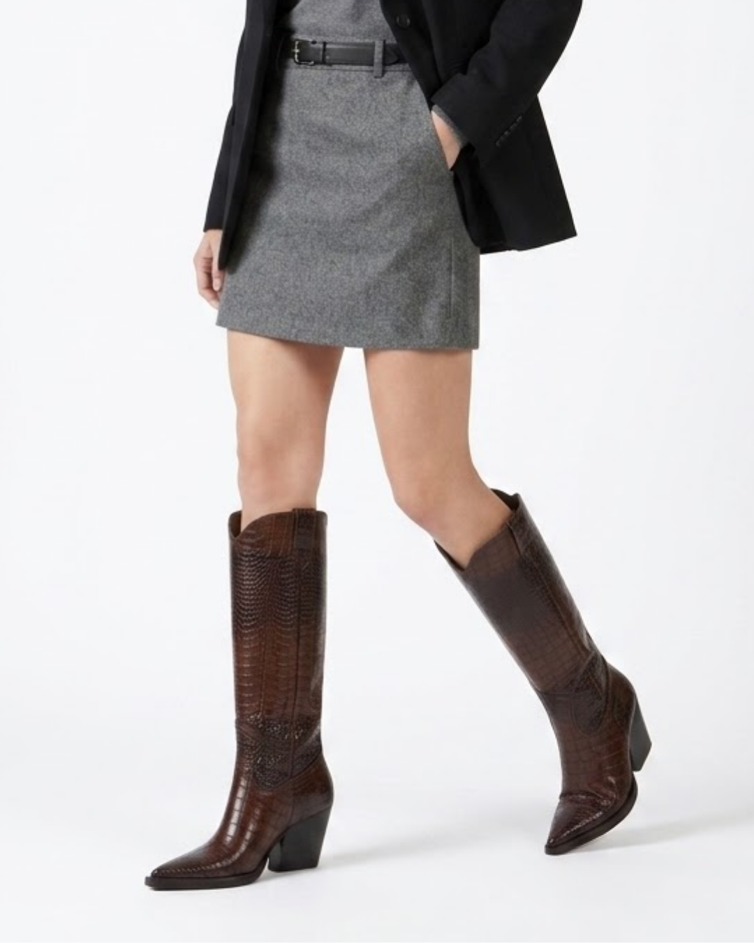 A model wearing a pair of dark brown textured knee-high cowboy boots, styled with a gray mini skirt, black blazer, and black belt against a plain white studio background.