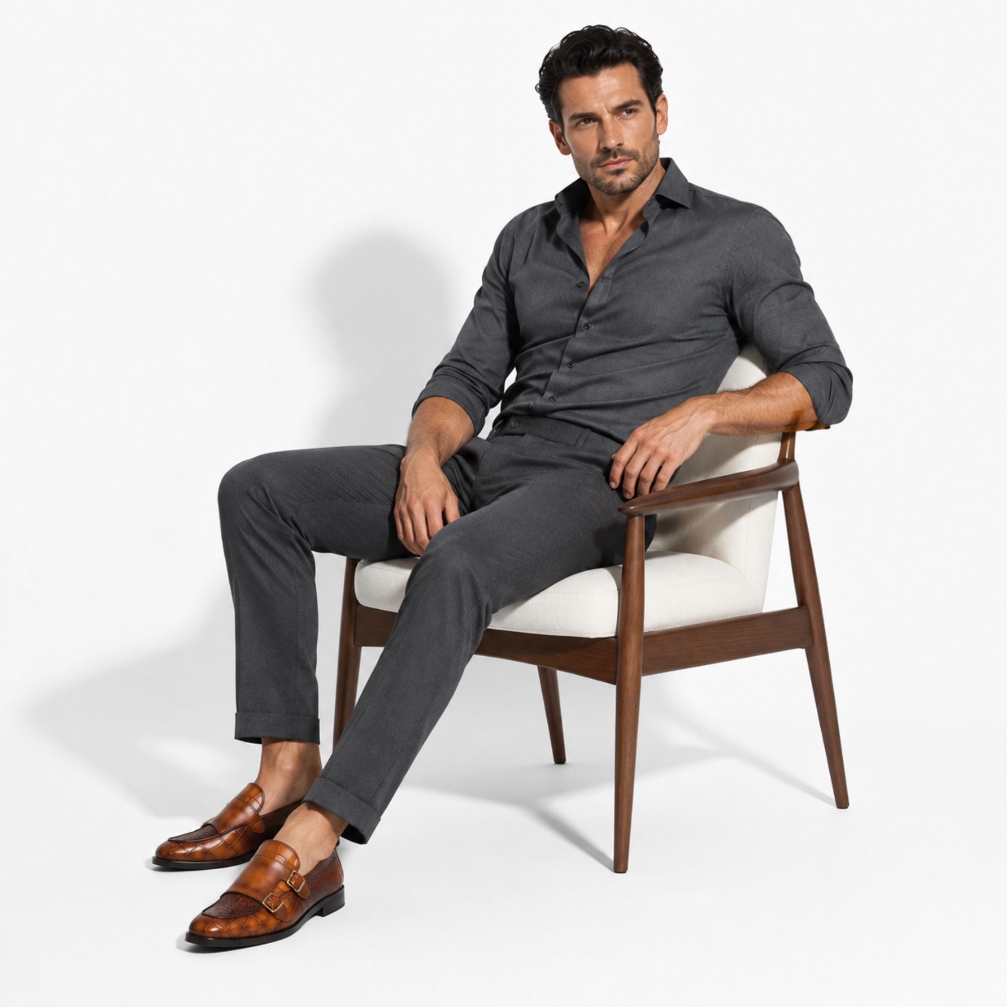 A man wearing a dark gray linen shirt and matching trousers sits in a modern wooden armchair with a white cushion, wearing brown leather loafers, against a clean white studio background.