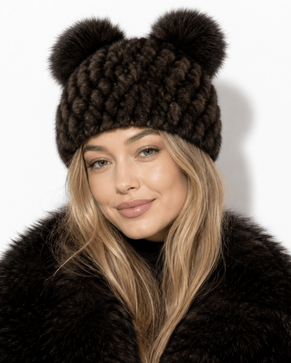 A woman wearing a dark brown chunky knit beanie with two large pom-poms on top, paired with a matching dark fur coat against a plain white background.