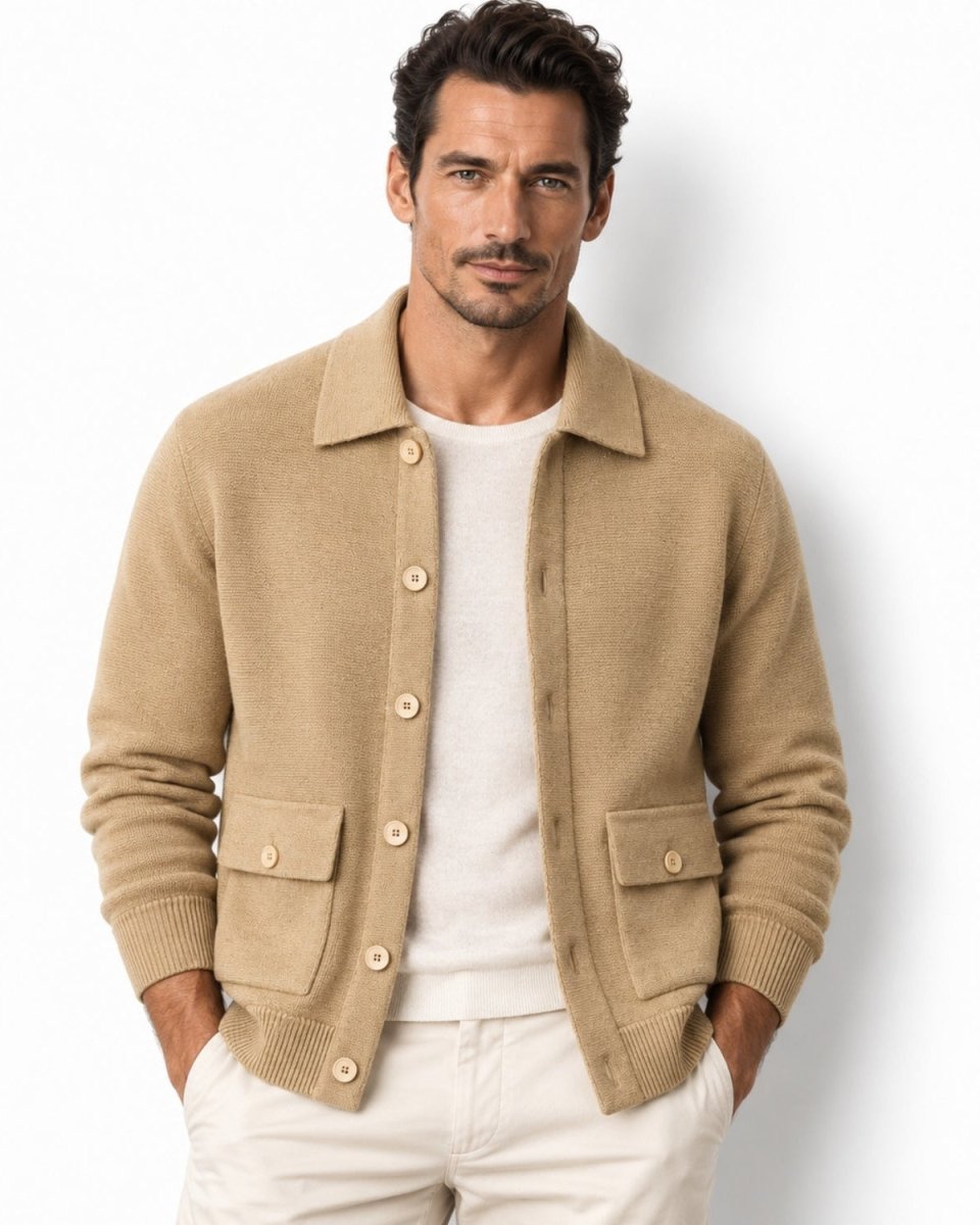 A man wearing a camel knit button-front cardigan over a white crewneck sweater and white pants, standing against a plain white studio background.
