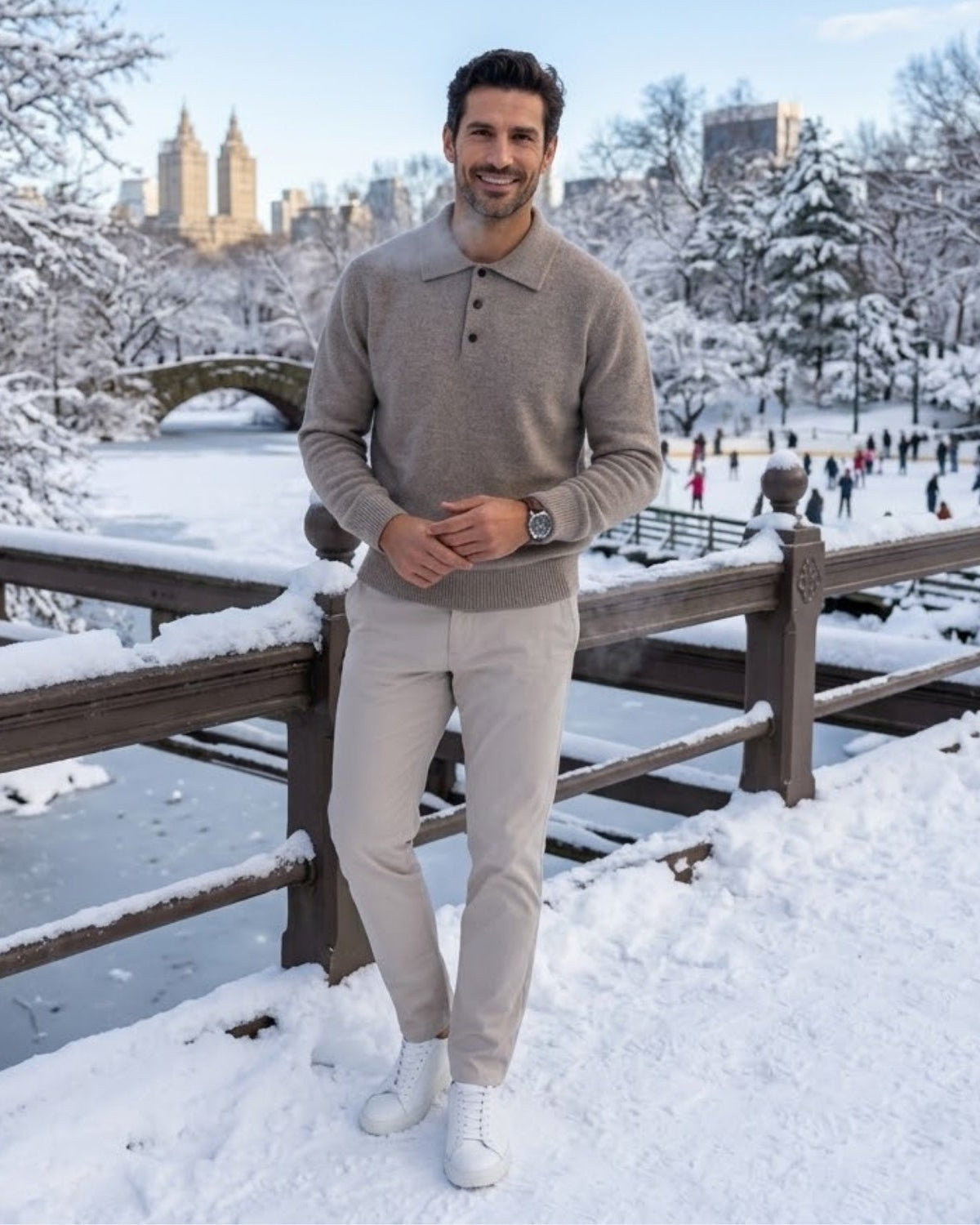 A man stands on a snowy bridge in Central Park, wearing a taupe knit polo sweater, light beige trousers, and white sneakers. The background shows a winter park scene with snow-covered trees, a frozen river, and city buildings in the distance.