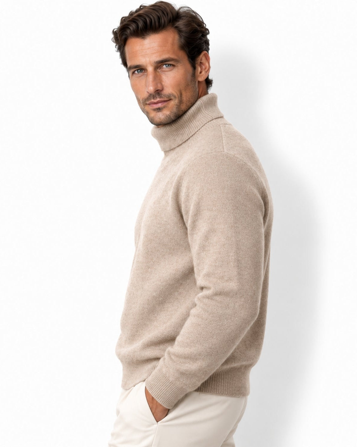 A man wearing a light camel knit turtleneck sweater and cream trousers, photographed against a clean white studio background.