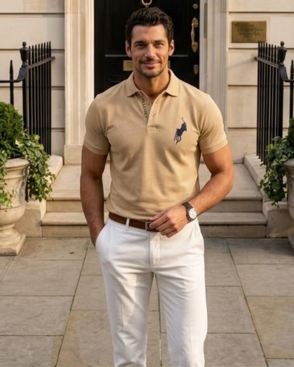 A man wearing a beige short-sleeve polo shirt with a navy embroidered polo player logo on the chest, paired with white trousers, standing in front of a classic stone building entrance.