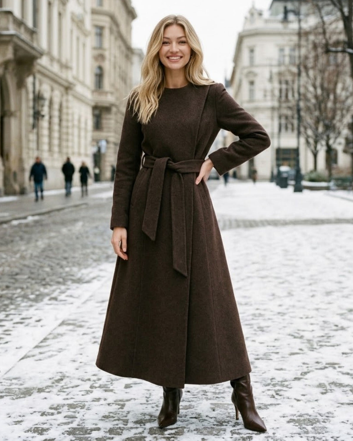 A woman stands on a snowy city street wearing a long, dark brown wrap-style coat with a matching waist tie and dark brown ankle boots.