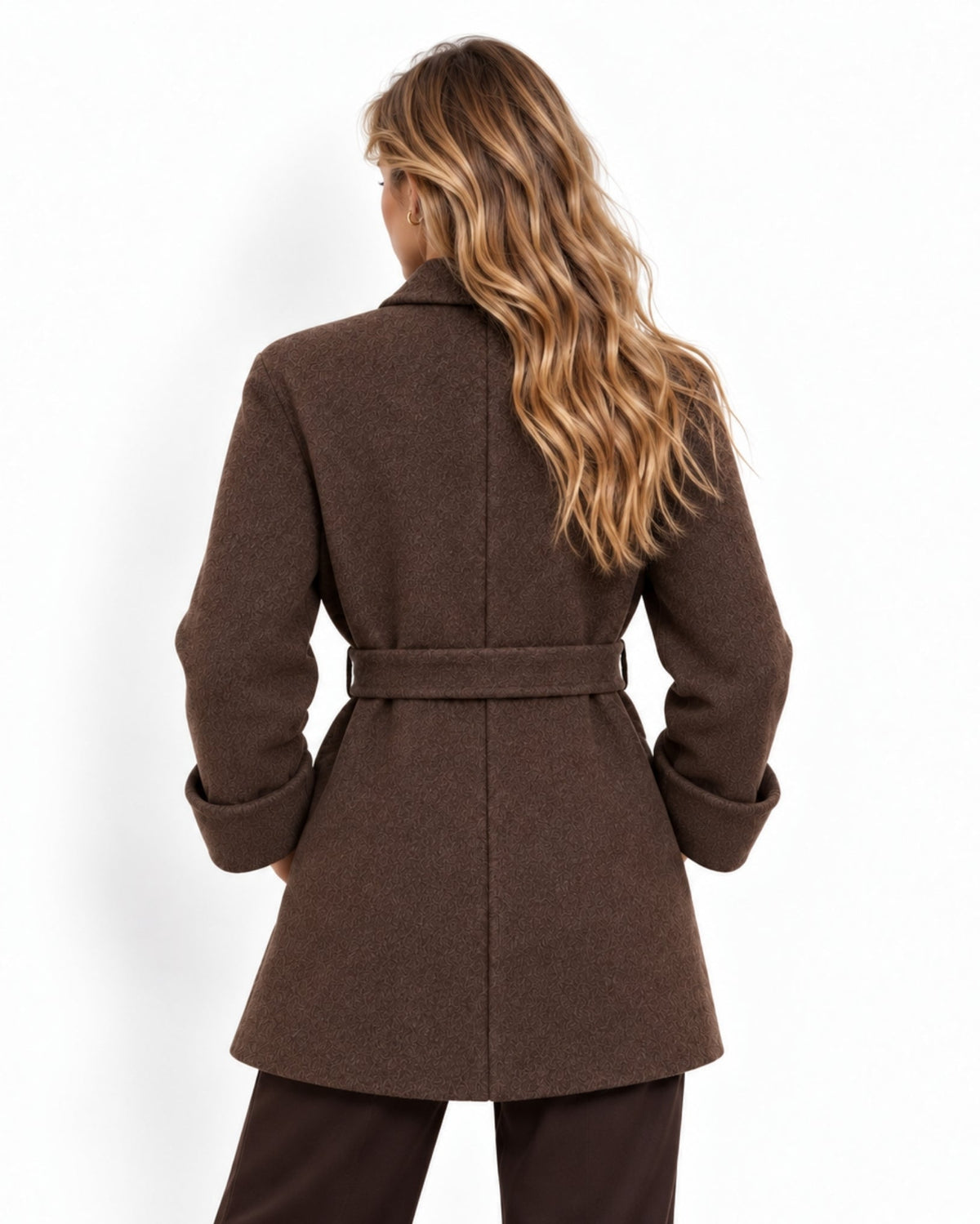 A woman viewed from the back wearing a brown belted coat and dark brown pants against a plain white studio background.