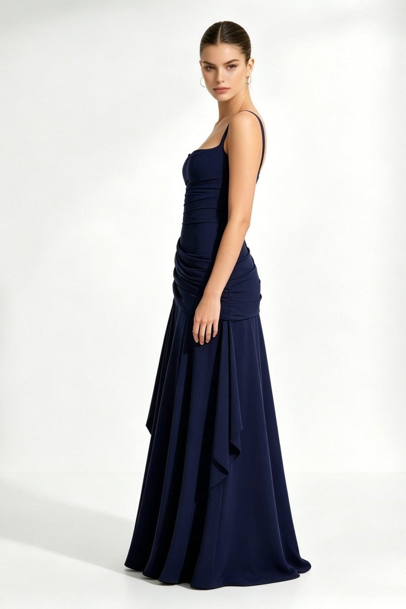 Side profile of a woman standing in a dark blue floor-length flowy formal maxi dress featuring structured asymmetrical draped hip details.