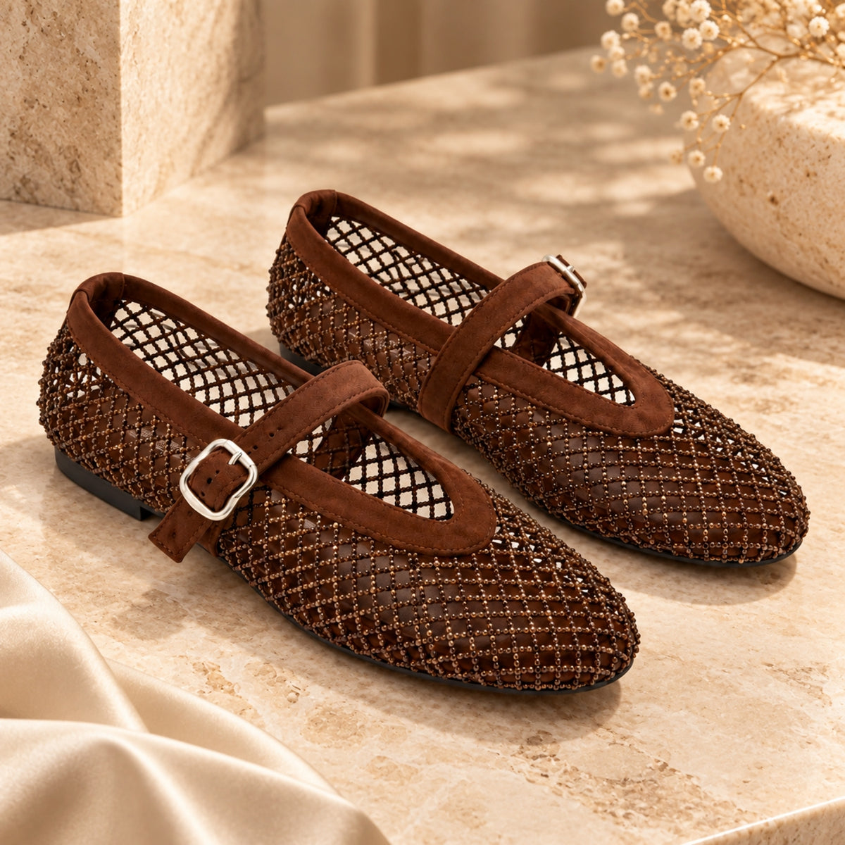 A pair of women's Mary Jane flats in brown, featuring a textured openwork mesh upper, a medium brown strap, and a silver buckle, displayed on a beige stone surface.