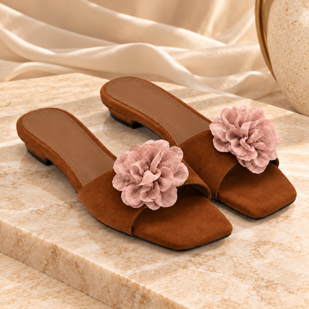 A pair of brown suede mule sandals with large dusty pink fabric flowers on the toe area, displayed on a beige marble surface with draped cream fabric in the background.