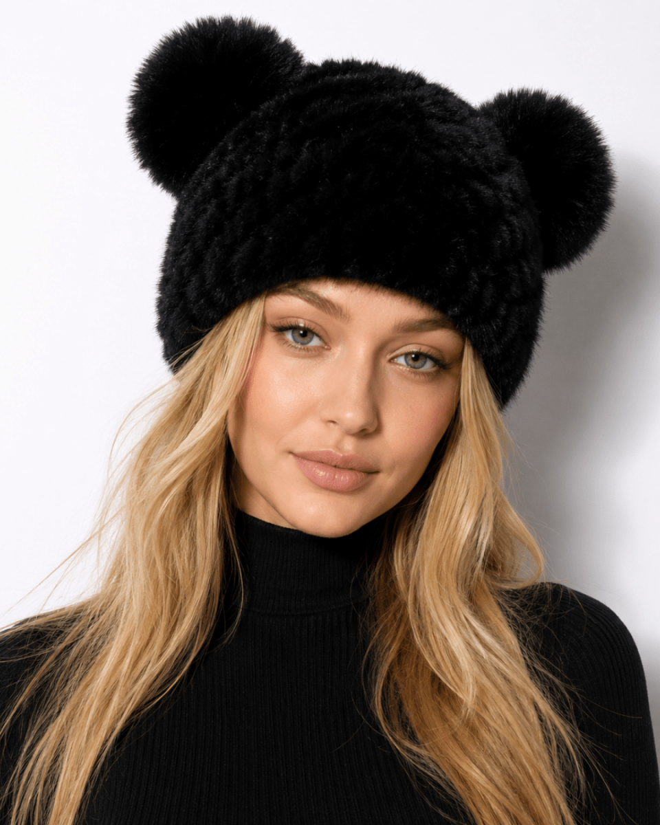 A woman with long blonde hair wearing a black faux fur beanie with two pom-pom ears, paired with a black ribbed turtleneck sweater, against a plain white background.