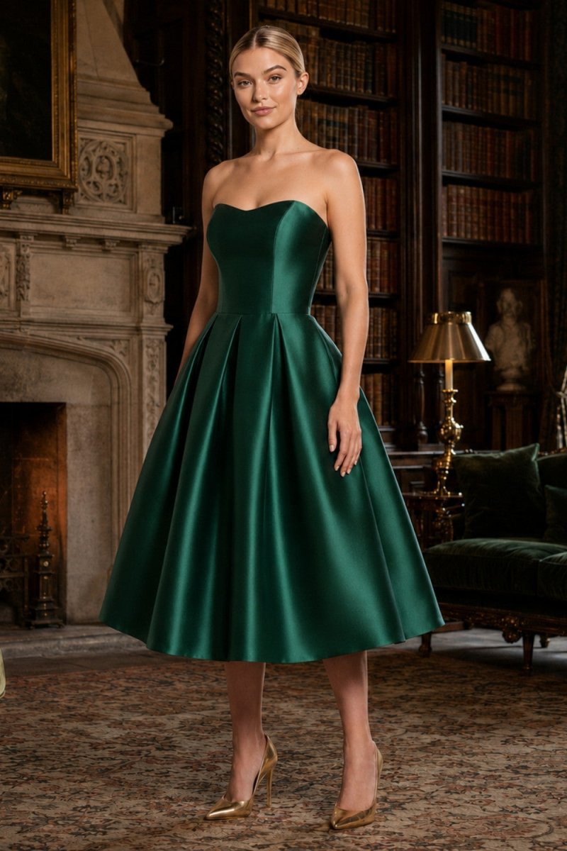 A woman modeling a strapless emerald green satin midi dress with a sweetheart neckline and full skirt, standing in a classic library room with bookshelves and a fireplace.