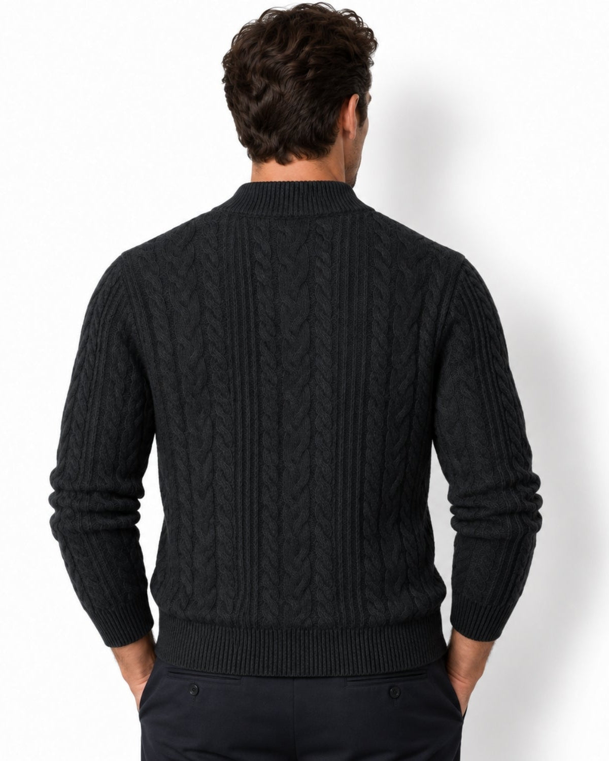 A man viewed from the dark grey wearing a black cable-knit sweater and dark trousers against a plain white studio background.