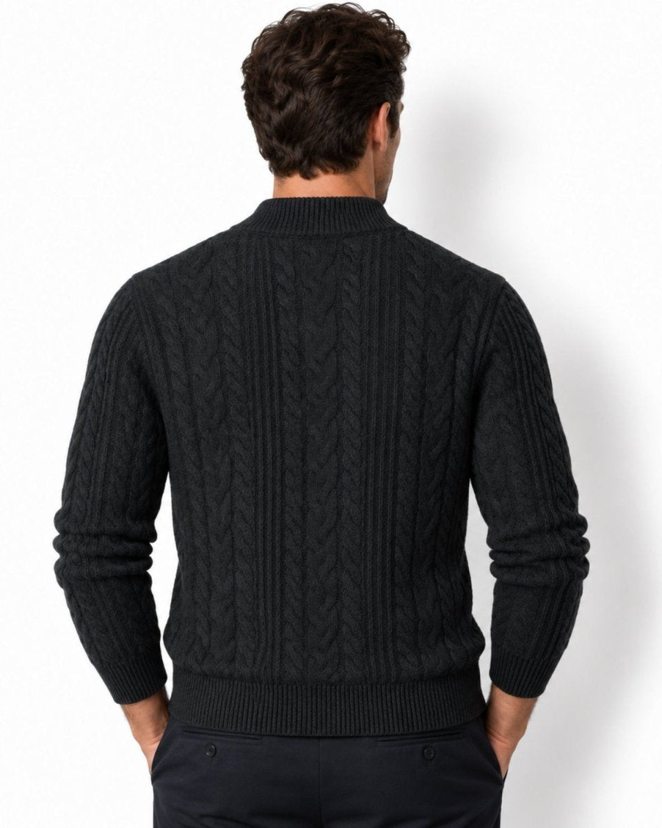 A man viewed from the dark grey wearing a black cable-knit sweater and dark trousers against a plain white studio background.