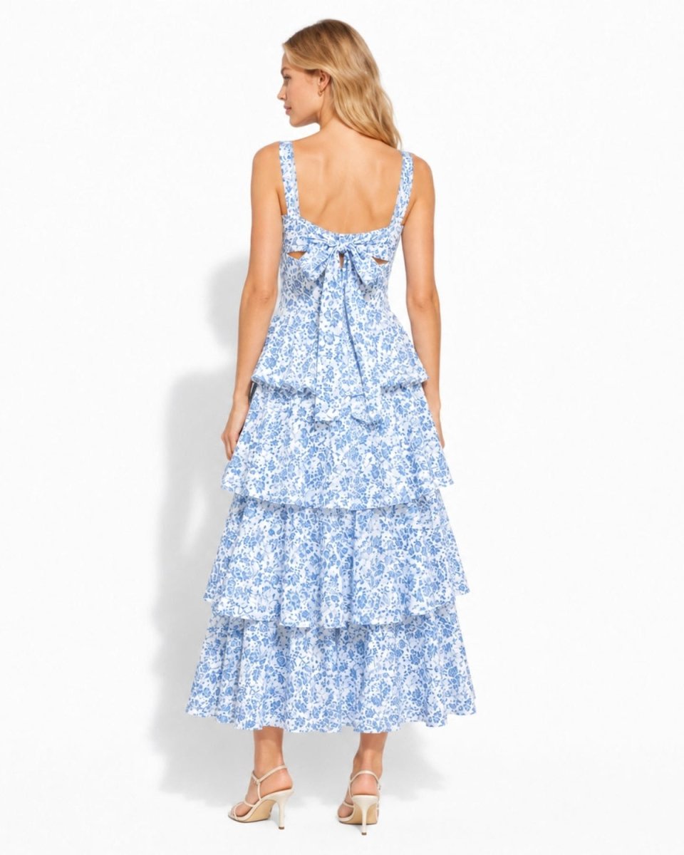 A woman modeling a sleeveless, tiered maxi dress from the back, featuring a blue and white floral pattern and a bow at the back neckline, set against a plain white studio background.