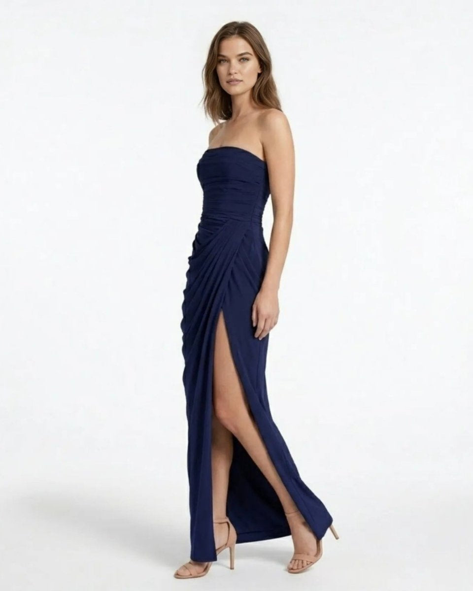 Side profile of a woman modeling a sophisticated navy blue strapless evening gown featuring dramatic draping and a high side slit.