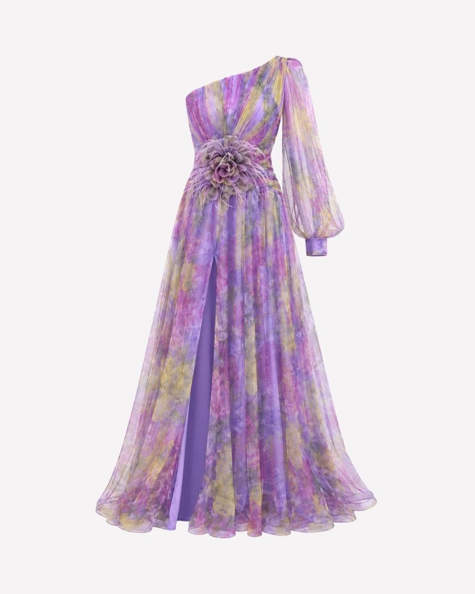 A one-shoulder floor-length gown with a purple, lavender, and yellow tie-dye pattern, featuring a large fabric flower detail at the waist and a sheer long sleeve, shown against a plain light gray background.