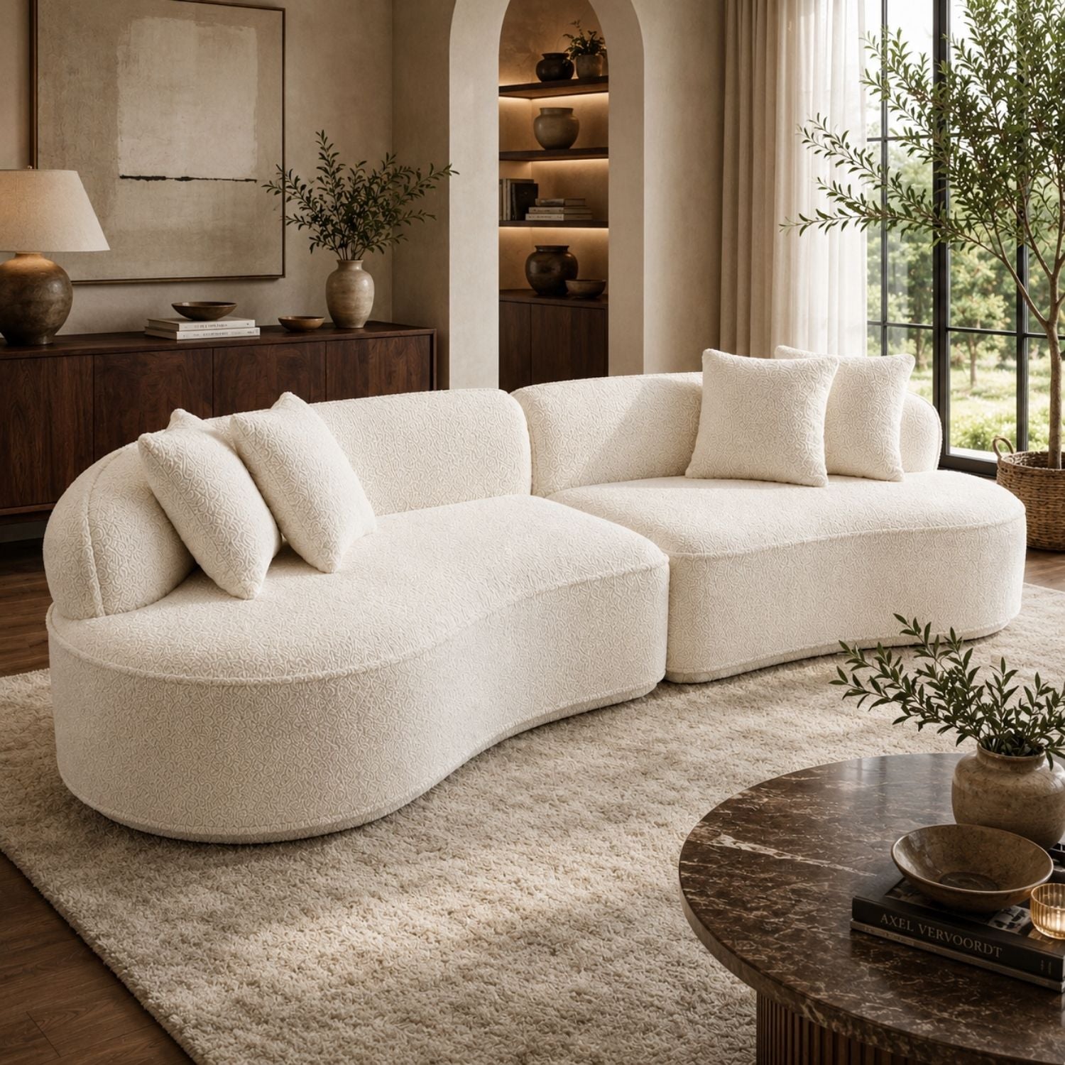 A cream-colored, curved modular sectional sofa with plush bouclé texture, styled in a warm, modern living room with a beige rug, wooden coffee table, and shelving.