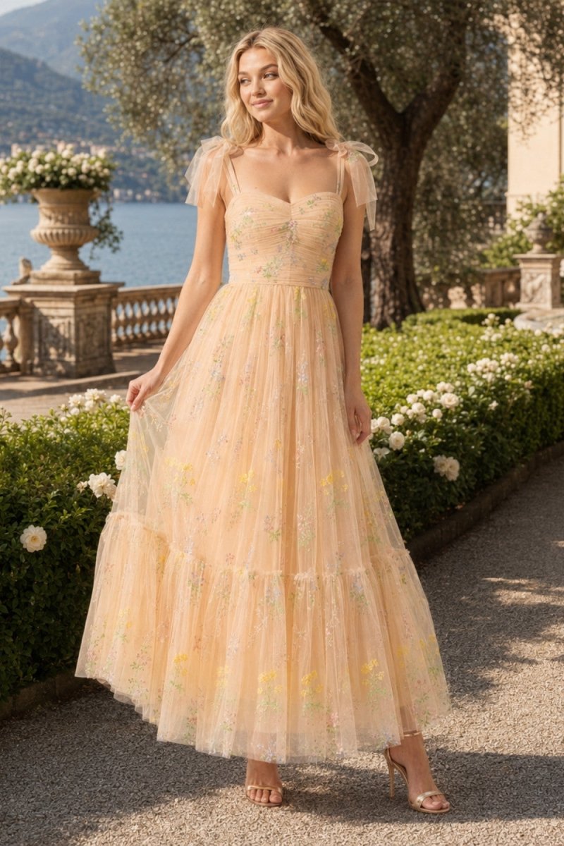 A woman wearing a flowing pale peach maxi dress with sheer flutter sleeves and embroidered floral details, standing outdoors on a garden path with flowers, trees, and water in the background.