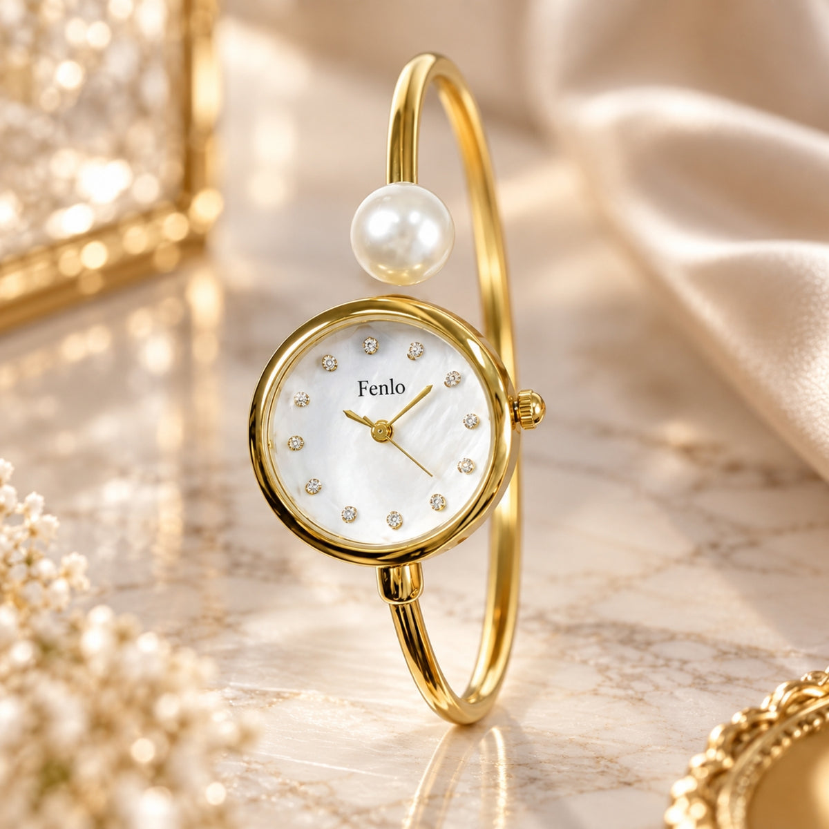A gold bangle watch with a round mother-of-pearl dial, crystal hour markers, and a single pearl accent on the upper curve, photographed on a soft beige marble surface with decorative gold and fabric props.