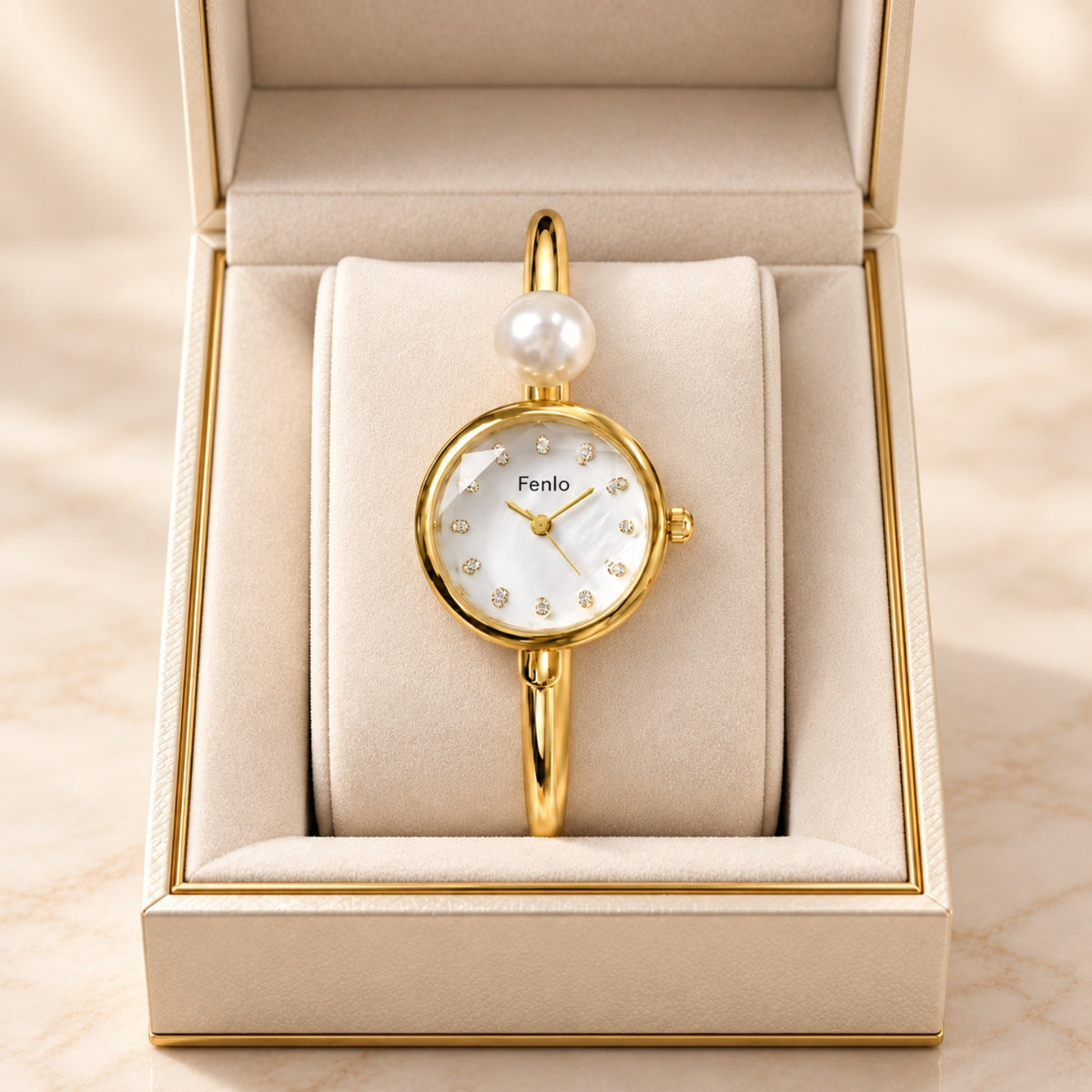 A gold-tone wristwatch with a white mother-of-pearl dial, crystal hour markers, and a single pearl accent at the top, presented inside an open cream-colored jewelry box.