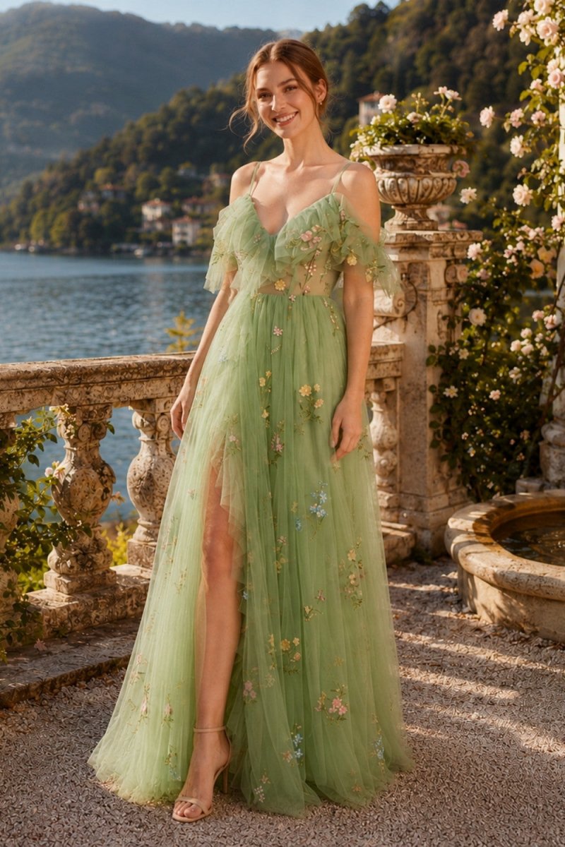 A woman stands on an outdoor terrace by a lake wearing a flowing off-the-shoulder green gown with floral embroidery and a high slit. Stone balustrades, plants, and water are visible in the background.