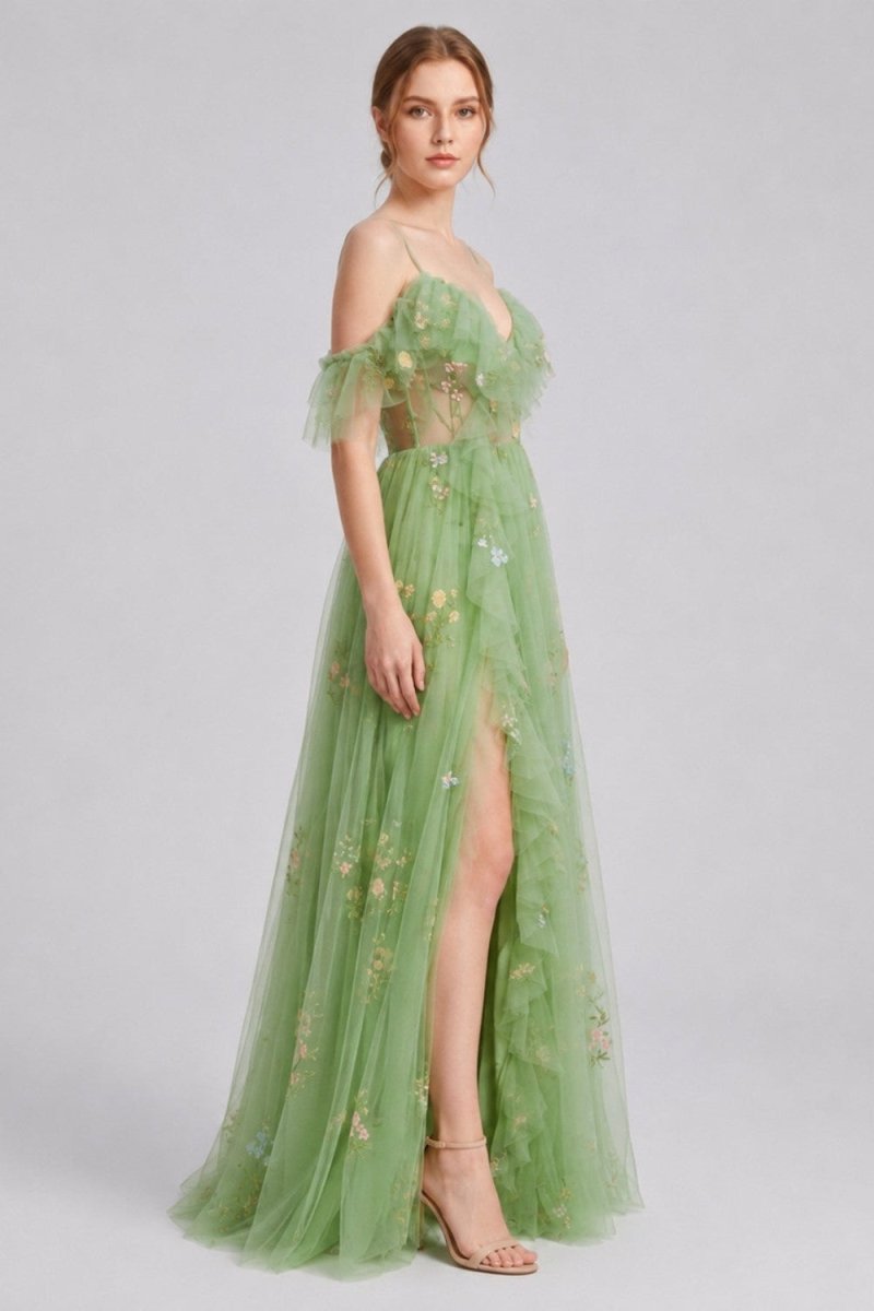 A woman models a floor-length sage green tulle gown with off-the-shoulder ruffled straps, a sheer embroidered bodice, and a high thigh slit, photographed against a light gray studio background.