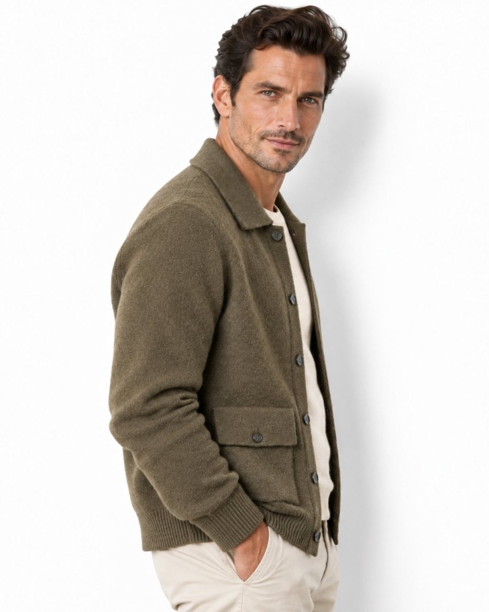 A man wearing an olive green knit button-up cardigan over a cream shirt and light beige pants, standing against a plain white studio background.