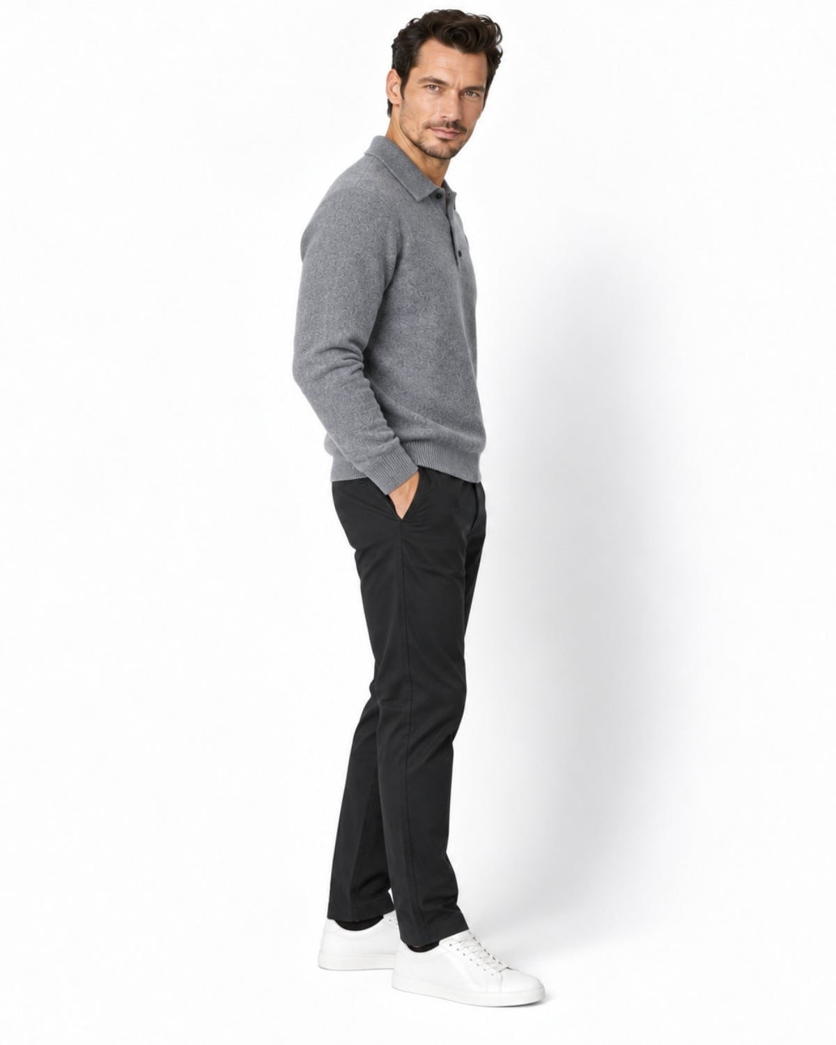 A man wearing a gray knit polo sweater, black trousers, and white sneakers, standing in profile against a clean white studio background.