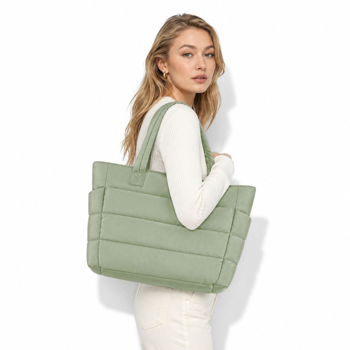 A woman wearing a white ribbed long-sleeve top and white pants carries a large sage green puffer tote bag against a clean white background.