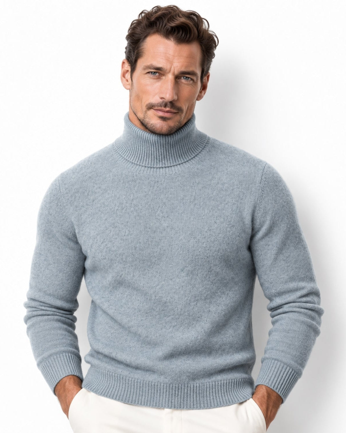 A man wearing a green ribbed turtleneck sweater and white trousers, standing against a plain white background.