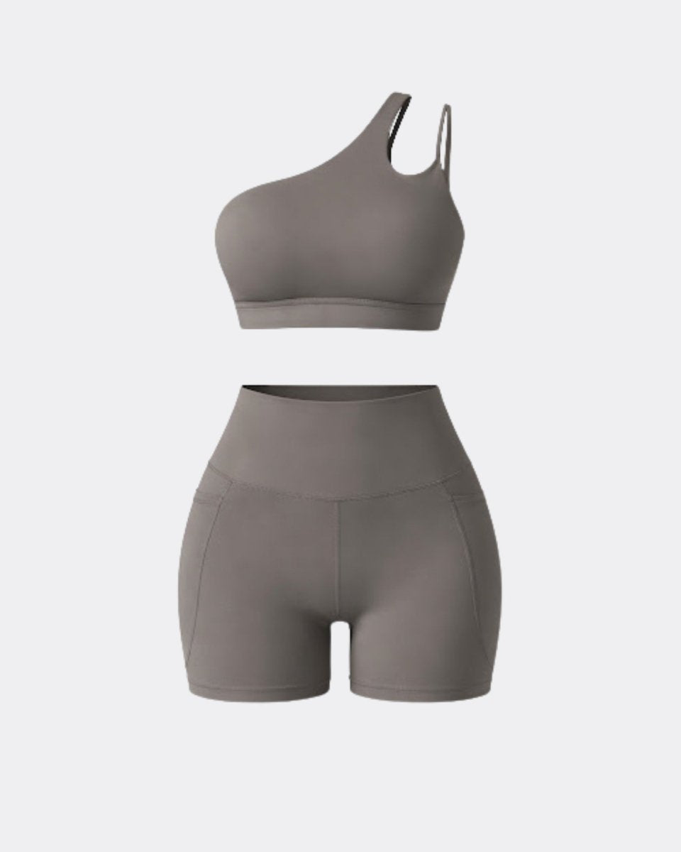 Flat lay studio shot of grey two piece workout outfit featuring asymmetrical top and pocket biker shorts.
