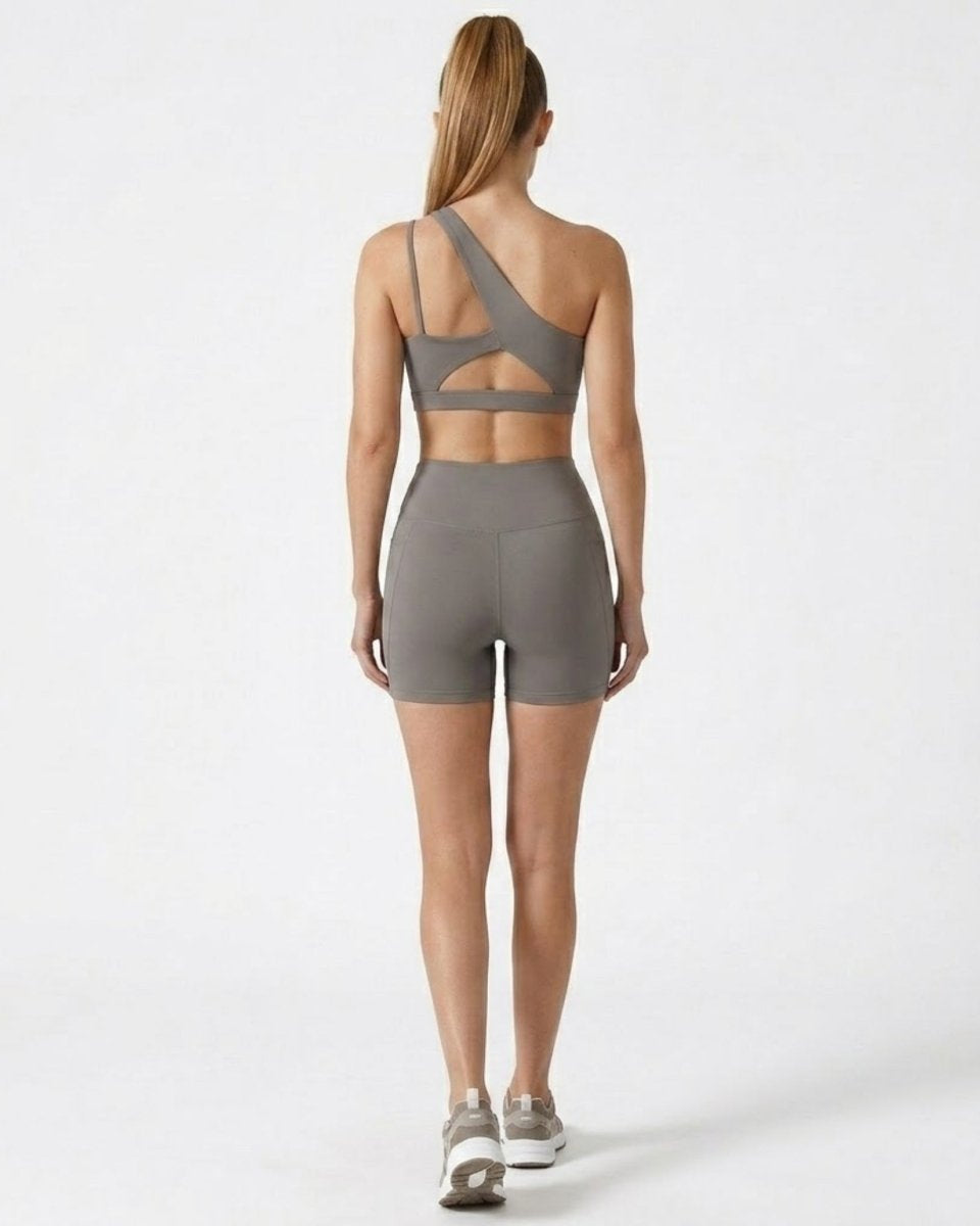 Back view highlighting the cutout strap detailing on the grey one shoulder sports bra and sculpting athletic shorts.