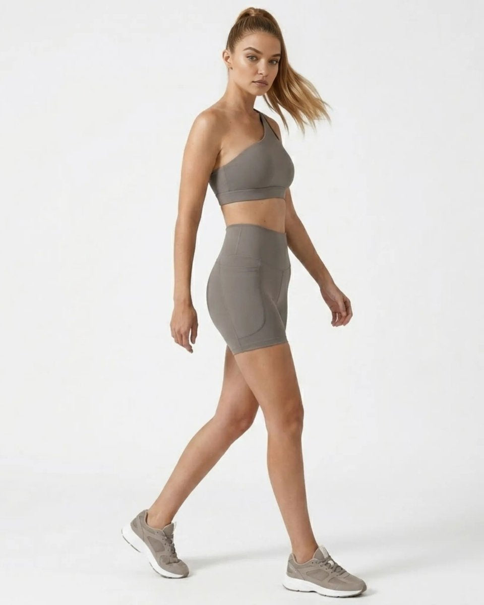 Side profile of model walking in grey high waisted workout shorts with side pockets and matching one shoulder top.