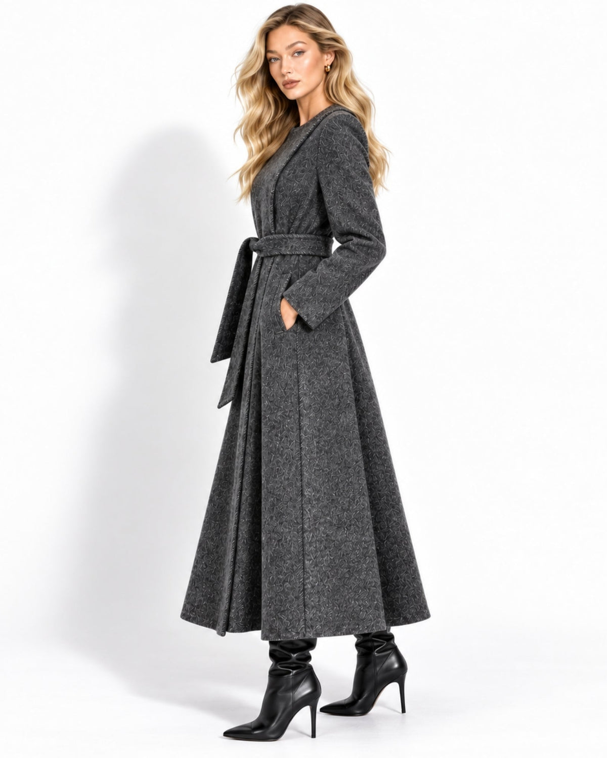 A woman modeling a long, dark gray belted wrap coat with a flared skirt, paired with black heeled ankle boots, against a plain white studio background.